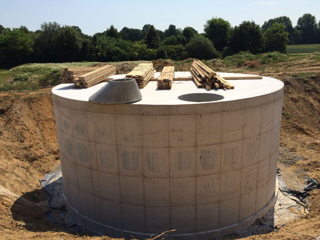 Runder Betonbehälter einer Kläranlage auf Baustelle, umgeben von Erde und Baumaschinen.